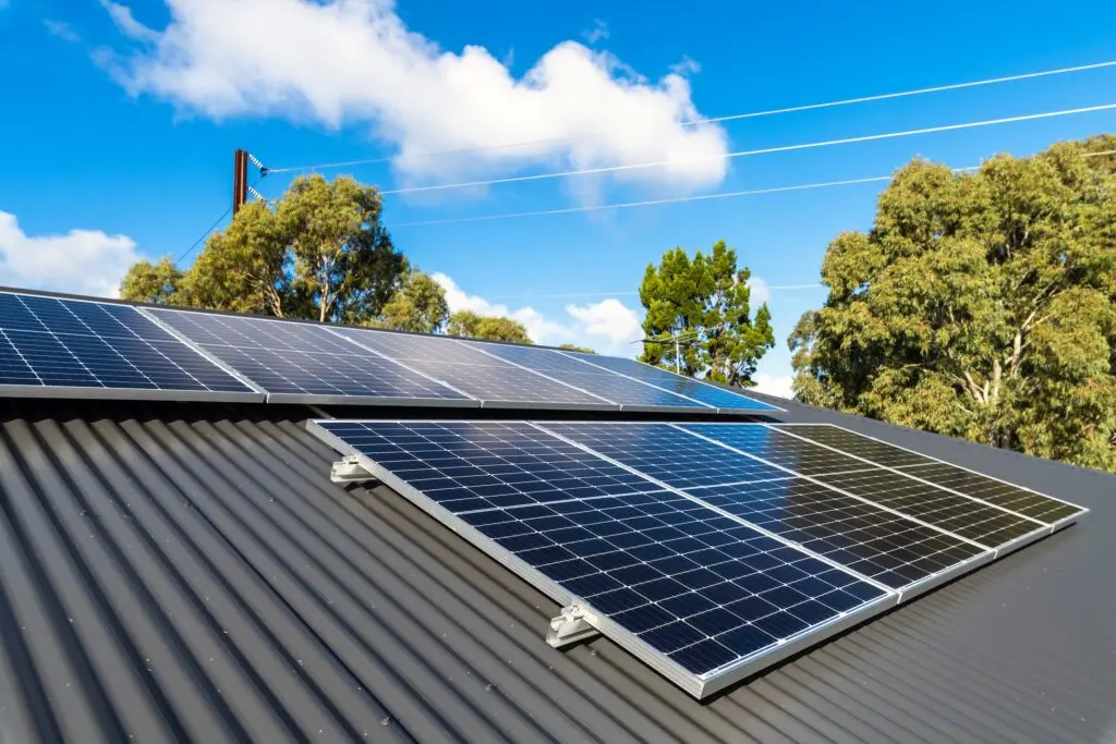 Solar panels on metal roof absorb sunlight atop a dark corrugated surface, framed by green trees and a blue sky with clouds in the background. Power lines stretch above the treetops, completing the scene.