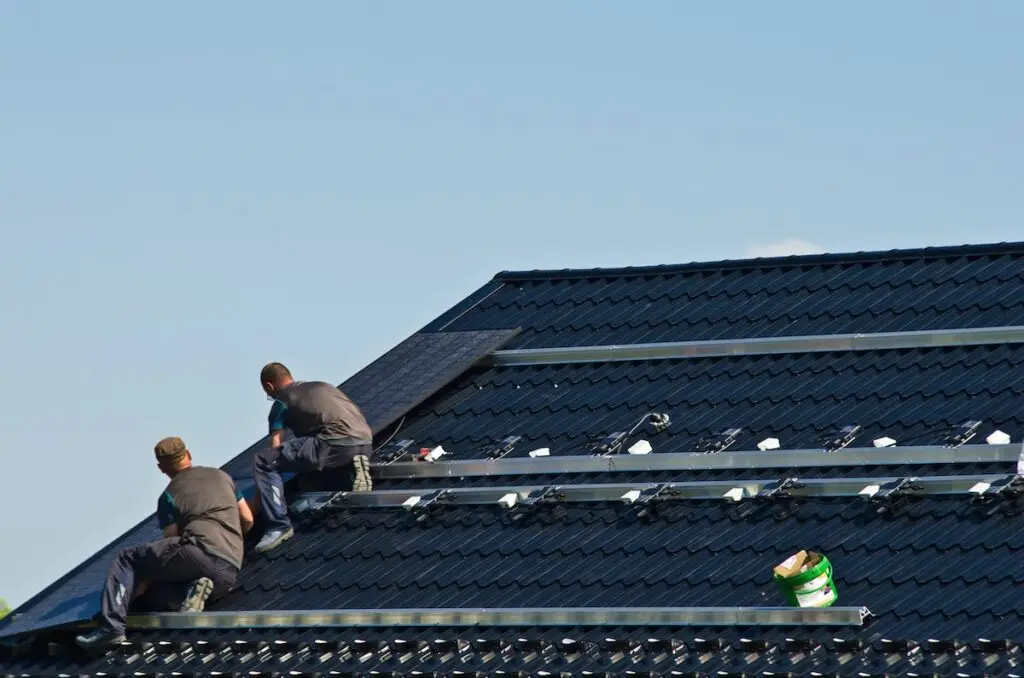 Two workers focus intently on a solar installation in Stoughton, fitting panels onto a dark, sloped roof beneath a clear blue sky. Dressed in gray outfits, they work with precision, one perched higher than the other. A green and white bucket sits nearby, completing the scene.