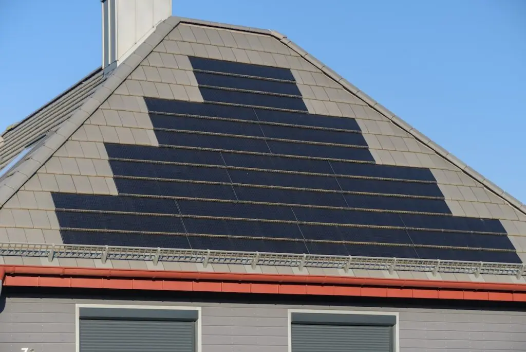 A triangular roof with solar panels arranged in a pyramid-like design against a clear blue sky. The roof has light-colored tiles, and two windows are partially visible below.