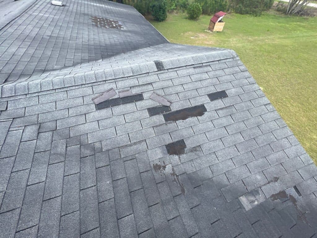Gray shingled roof with several missing and damaged shingles, exposing dark patches underneath. The lawn and some trees are visible in the background along with a small wooden structure.