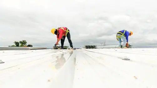 commercial roof repair services Two construction workers wearing safety gear and harnesses are working on a white metal roof under a cloudy sky. One is crouched, using tools, while the other is bent over, both focused on their tasks.