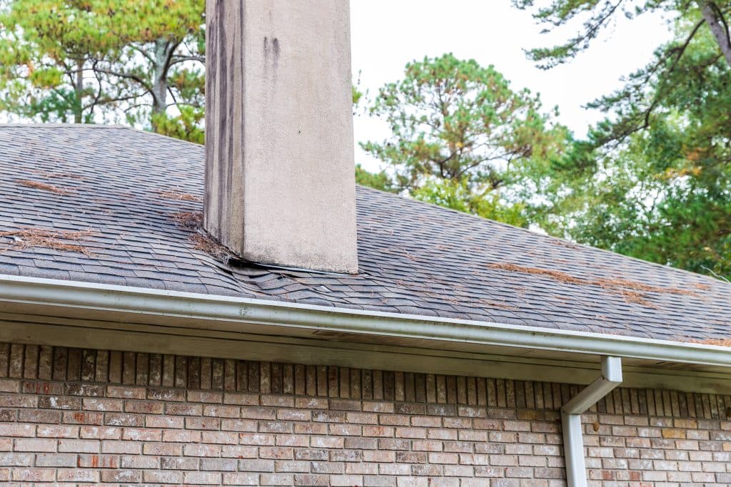 A brick house with a shingled roof shows water damage and sagging near a chimney, with pine needles scattered on the roof and trees in the background.