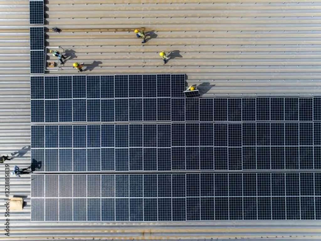 Aerial view of workers in yellow helmets diligently performing a solar installation in Fithburg on a large, ribbed metal roof. The panels cover a significant portion of the roof, reflecting sunlight, with workers strategically positioned in the upper section.