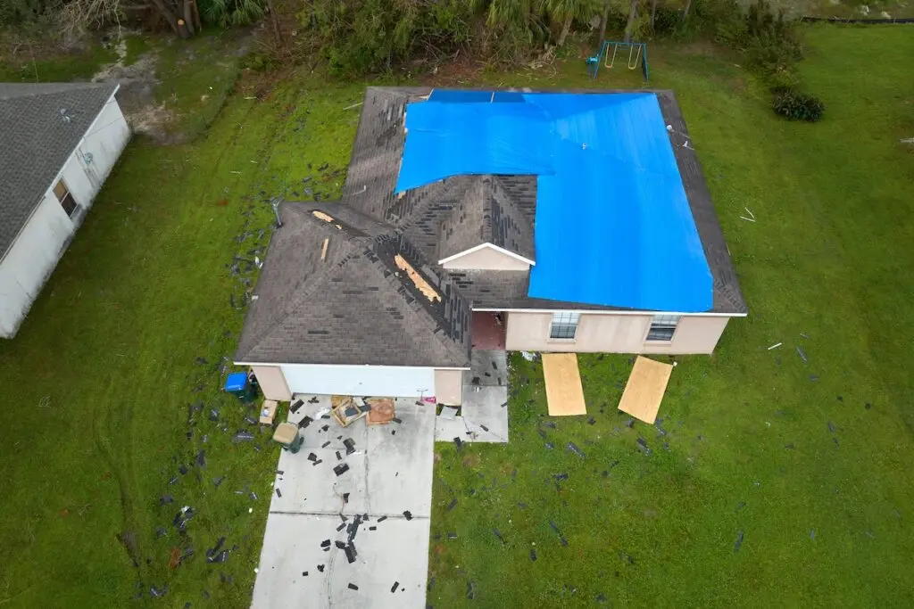 Aerial view of a house with a blue tarp covering part of the roof, indicating damage. Shingles and debris are scattered across the lawn and driveway. Two sheets of plywood lean against the front of the house. A swing set is in the backyard.