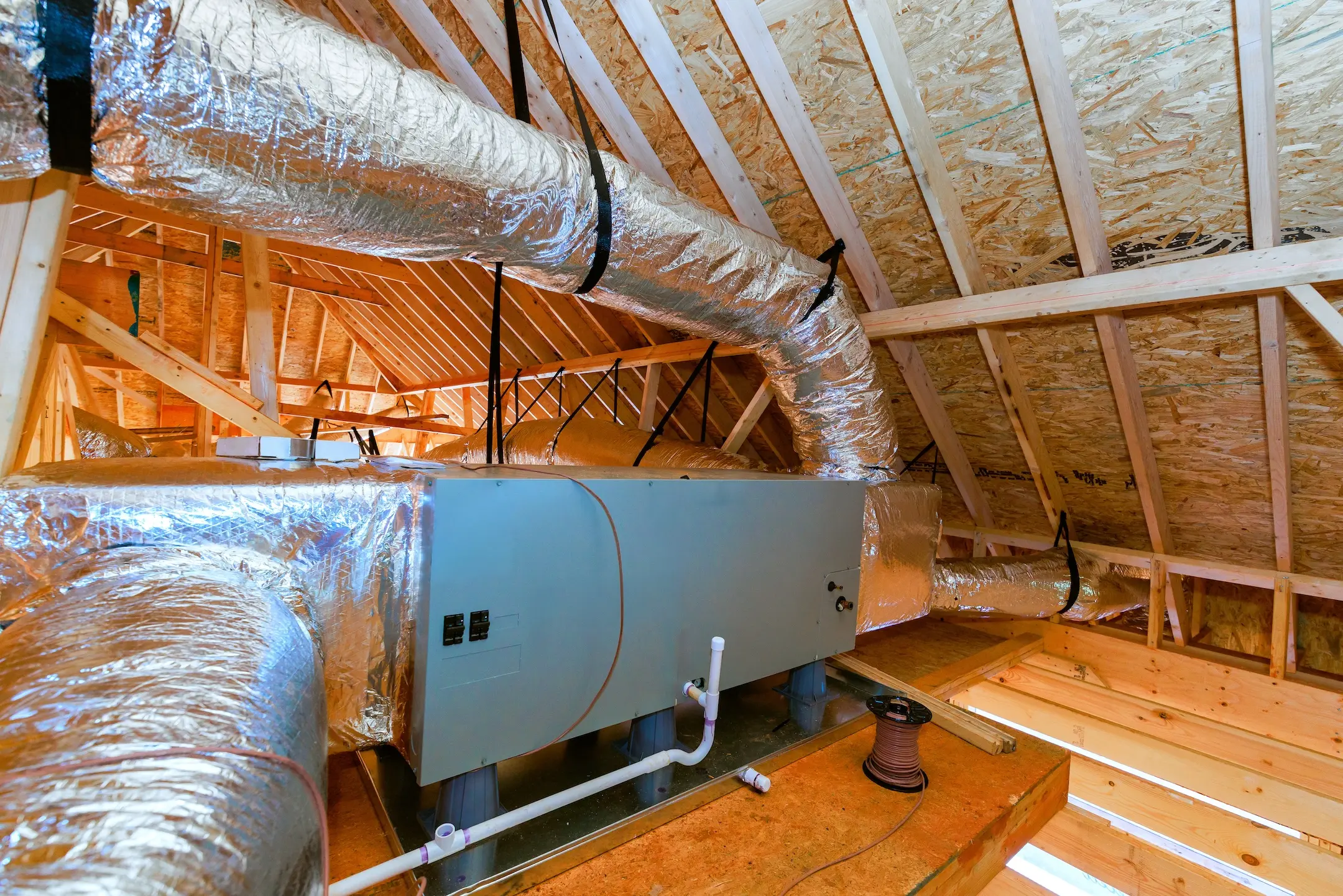 An HVAC unit with insulated ducts installed in an unfinished attic with exposed wooden beams and plywood walls.
