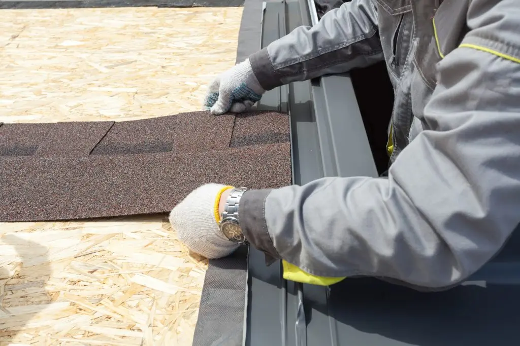 A person in work gloves and a gray jacket installs asphalt shingles on a wooden roof surface, preparing for roofing installation.