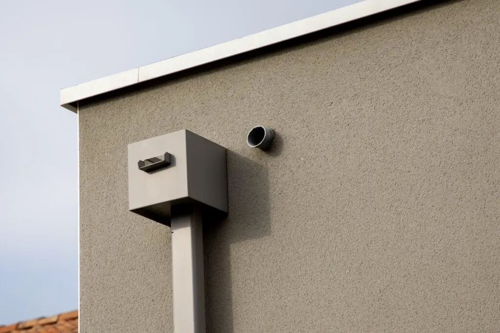 A close-up of a modern building’s exterior wall with a gray textured surface, featuring a rectangular metal vent and a circular pipe protruding from the wall.