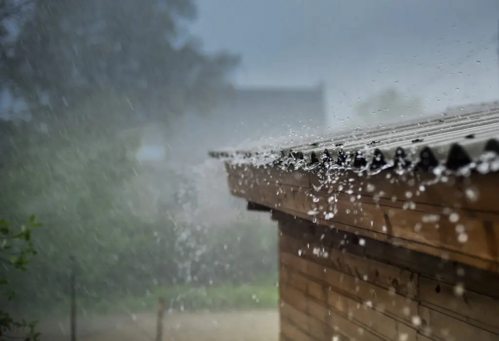 metal roof hail damage Heavy rain pours down on a corrugated metal roof of a wooden building, with water splashing off the edge and creating droplets in the air; a blurred, rainy landscape is visible in the background.