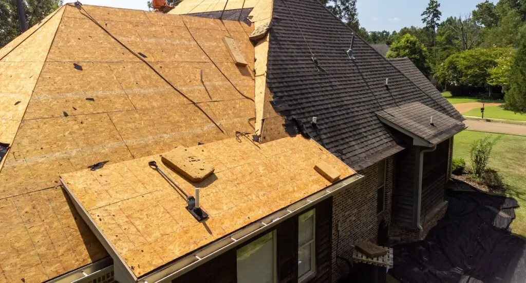 A house undergoing roof repair with sections of plywood exposed and shingles removed. Various tools are scattered on the plywood. Surrounding the house are trees and a lawn.