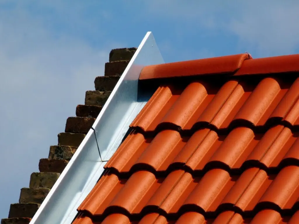 Close-up of a roof with red terracotta tiles, a metal flashing along the edge, and a section of brick chimney, under a partly cloudy blue sky.