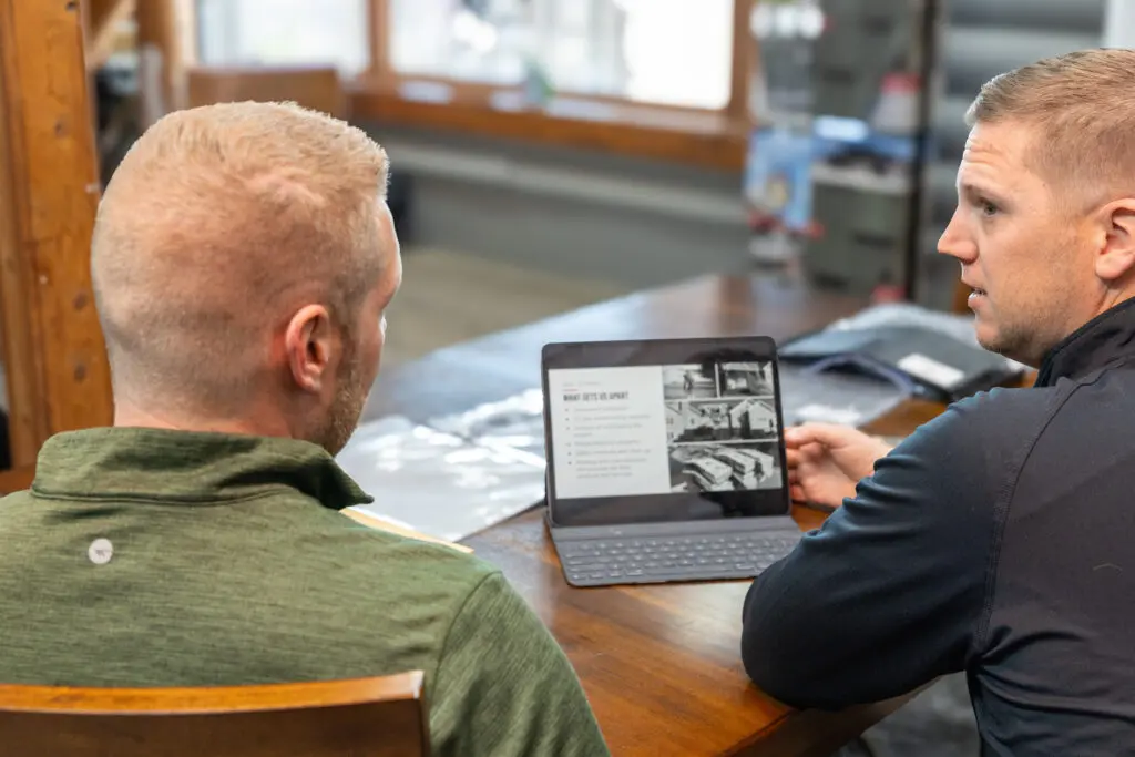 what wind speed will tear off a roof in stoughton wi Two men sit at a wooden table, one showing the other a laptop screen with text and black-and-white images. The setting appears to be a casual office or workspace with natural light from large windows.