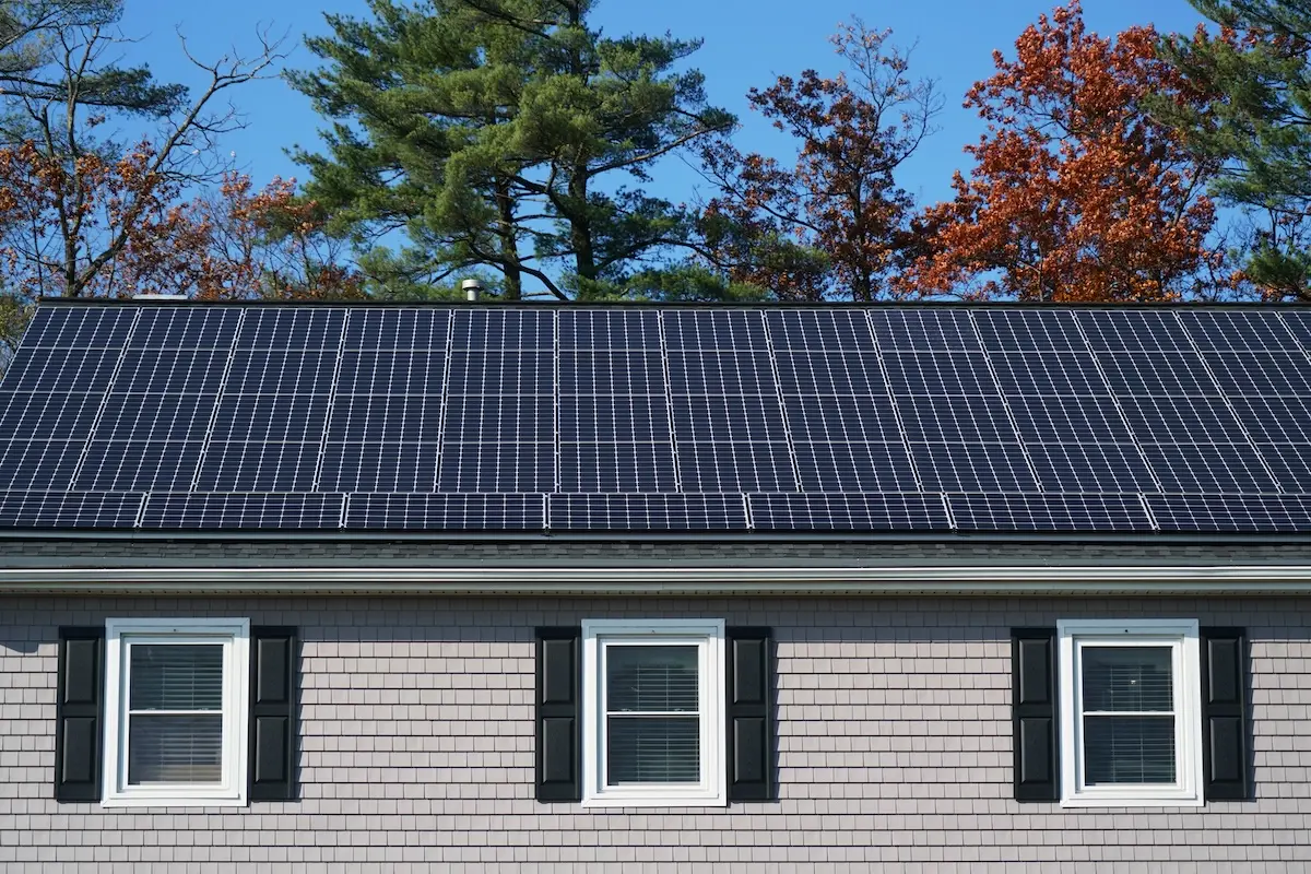 A house in Fitchburg boasts a solar installation covering the roof. The building features light-colored siding and three windows with black shutters. In the background, trees displaying green and autumn hues stand under a clear blue sky.