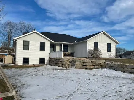 A white house with black trim sits behind a stone retaining wall. There is patchy snow on the ground, leafless trees in the background, and a blue sky with scattered clouds overhead.