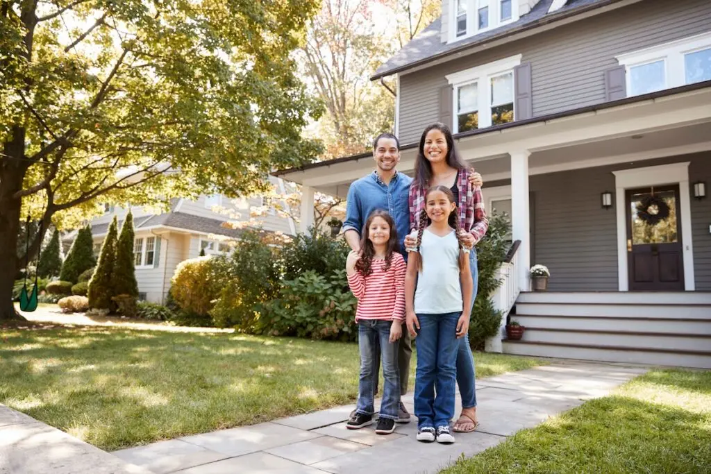 A smiling family of four stands on a walkway in front of their suburban house, recently enhanced by a solar installation. The parents are behind the two children, with green trees and a lawn surrounding the gray house in Waunakee as their backdrop.