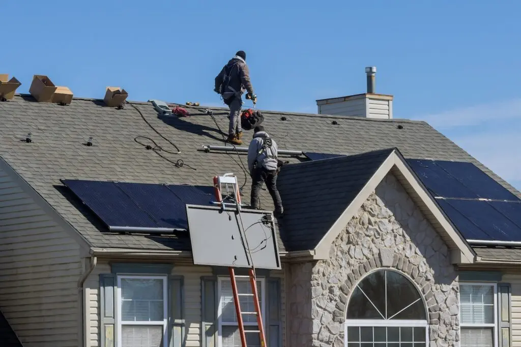 Two workers wearing safety harnesses install solar panels on the roof of a house. Ladders and equipment are on the roof, and several cardboard boxes are near the workers. The sky is clear and sunny.