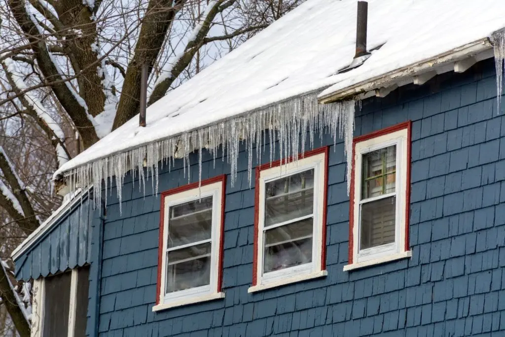 ice and water shield roof Blue house with red-trimmed windows, snow on the roof, and long icicles hanging from the eaves. Bare tree branches are visible in the background.
