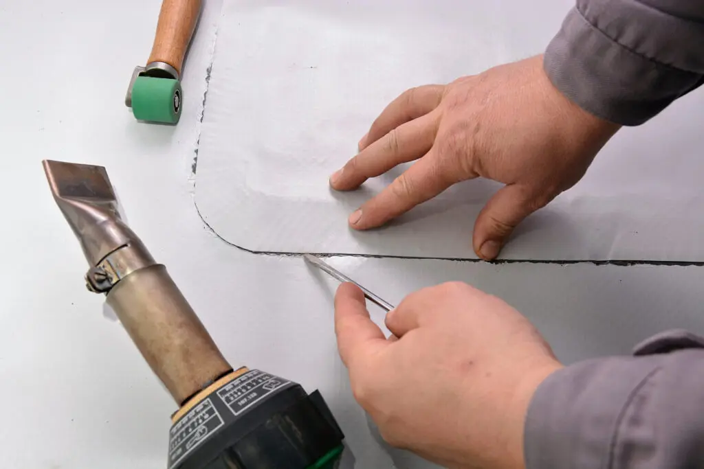 types of commercial roofing systems Hands using a utility knife to cut a sheet of material, with a hot air gun and a small roller tool nearby on a flat surface.