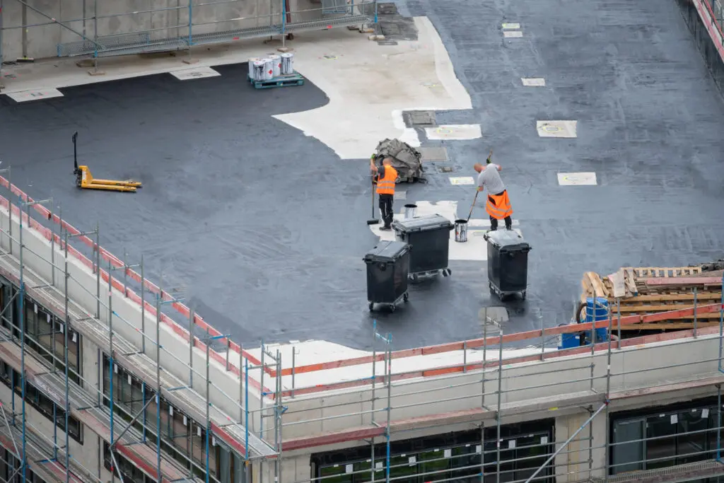 commercial roof repair services Two workers in orange vests handle large bins on a flat rooftop under construction, surrounded by scaffolding and building materials.