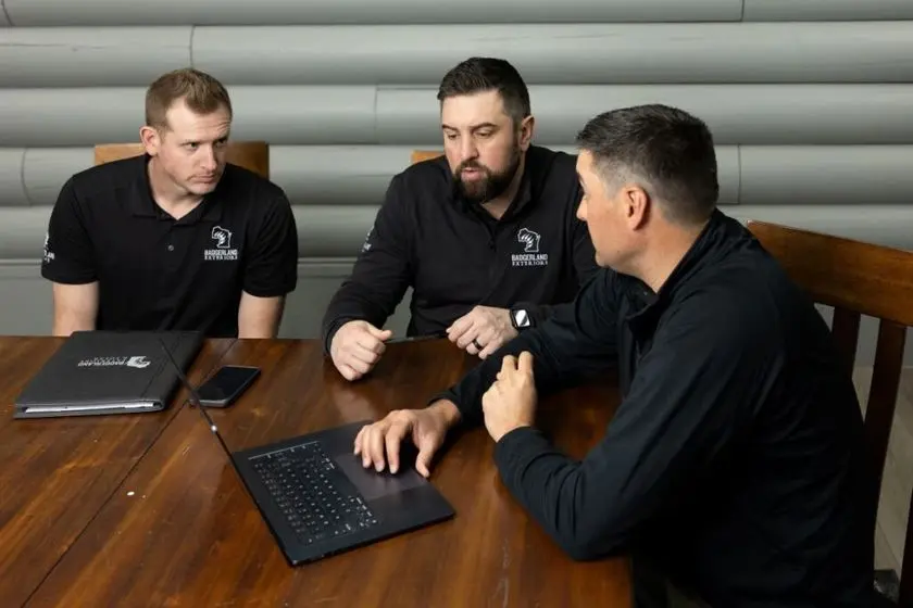 Three men sit around a wooden table, discussing something while looking at a laptop. One gestures with his hands, another listens closely, and a folder and phone are on the table. All wear black shirts with a white logo.