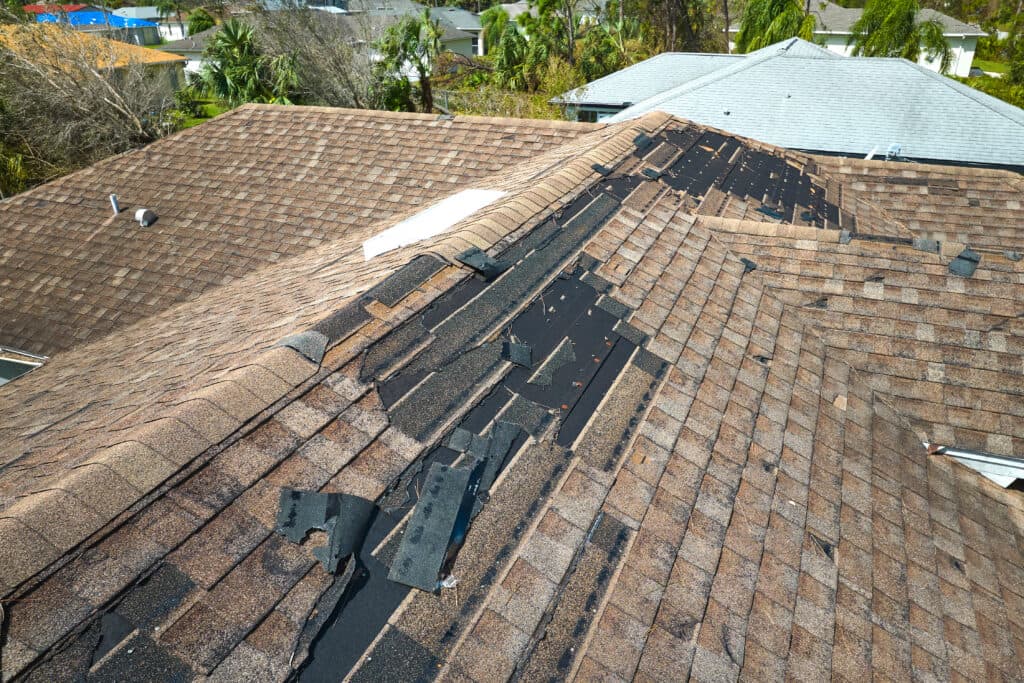 A house roof with brown shingles shows extensive storm damage, with missing shingles on roof exposing the black underlayment beneath. Nearby roofs appear intact.