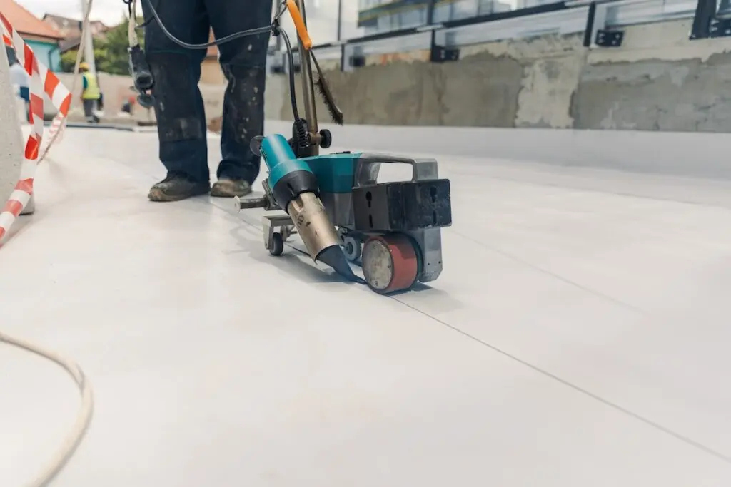 A worker uses a handheld welding machine to seal seams on a white synthetic flooring material at a construction site. Only the lower part of the worker and the equipment are visible.
