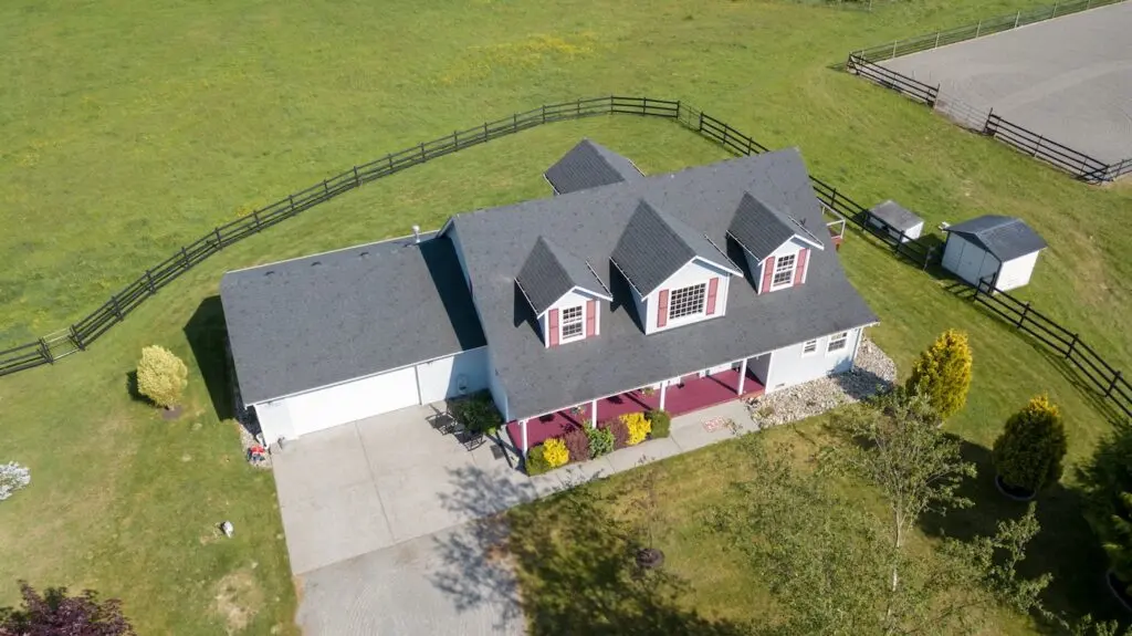 Aerial view of a two-story house with a gray roof, white trim, and a covered front porch—perfect for those learning how to measure a roof for shingles. The home features a large green lawn, fenced yard, and a small shed in the background.