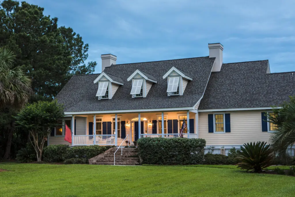 A large, two-story house with a grey shingled roof and three dormer windows. The house has white siding, a wraparound porch, and American flags near the entrance. Surrounded by greenery and palm trees, it's set against a blue sky.