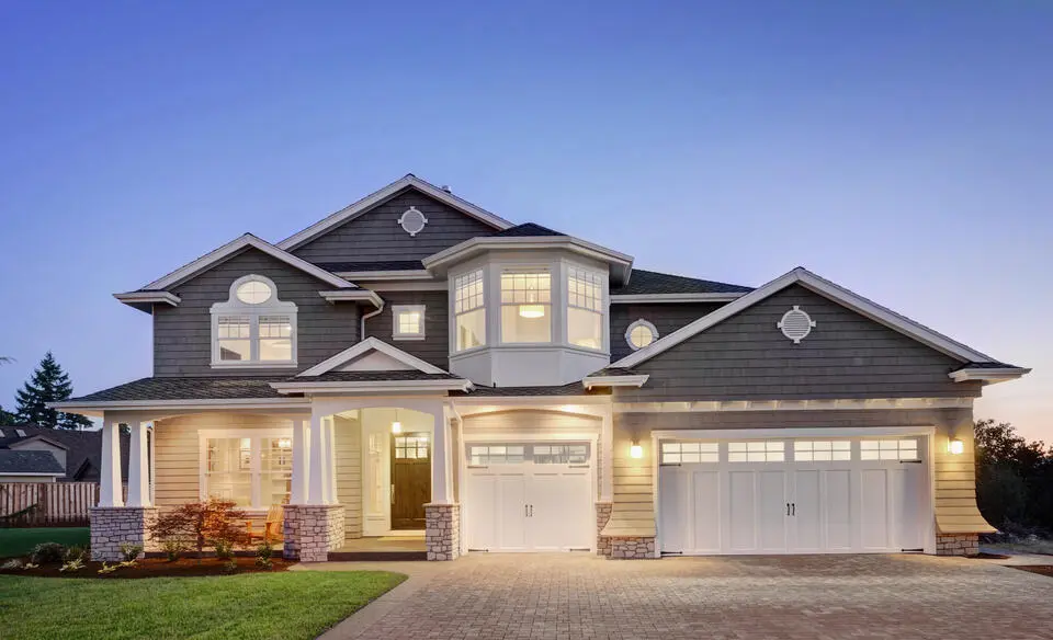 A two-story house with gray siding and white trim, featuring a large garage and multiple windows. The porch is lit warmly, and the sky is a gradient of blue, indicating dusk or dawn. Surrounding the house is a neatly landscaped garden.