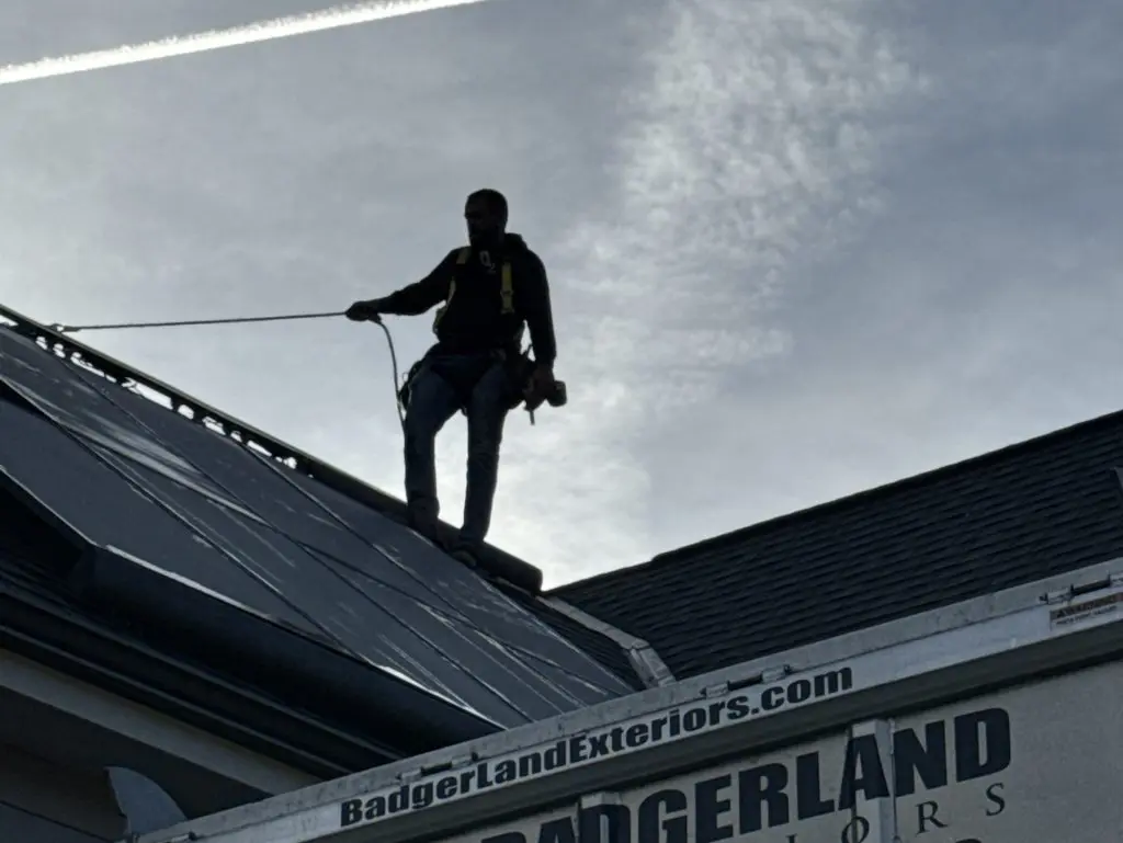 A worker wearing safety gear stands on a roof next to solar panels, secured by a harness. Below, a Badgerland Exteriors truck with the company’s phone number is visible. The sky is partly cloudy with a jet trail overhead.