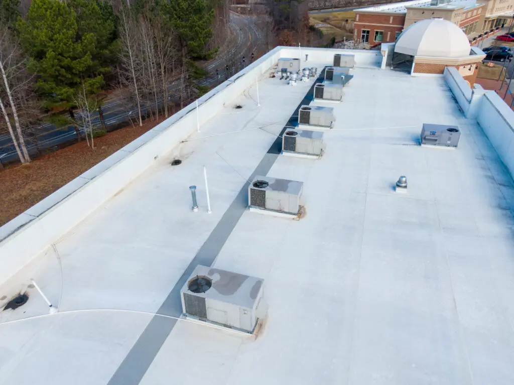 commercial roof repair services Aerial view of a flat, white commercial building roof with multiple HVAC units, surrounded by trees and a road, and a dome structure visible in the background.