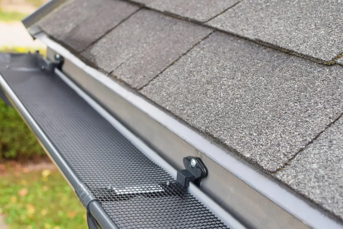 Close-up view of a house roof with asphalt shingles and a black mesh gutter guard installed along the edge, showcasing how to install gutter guards to prevent debris from entering the rain gutter.