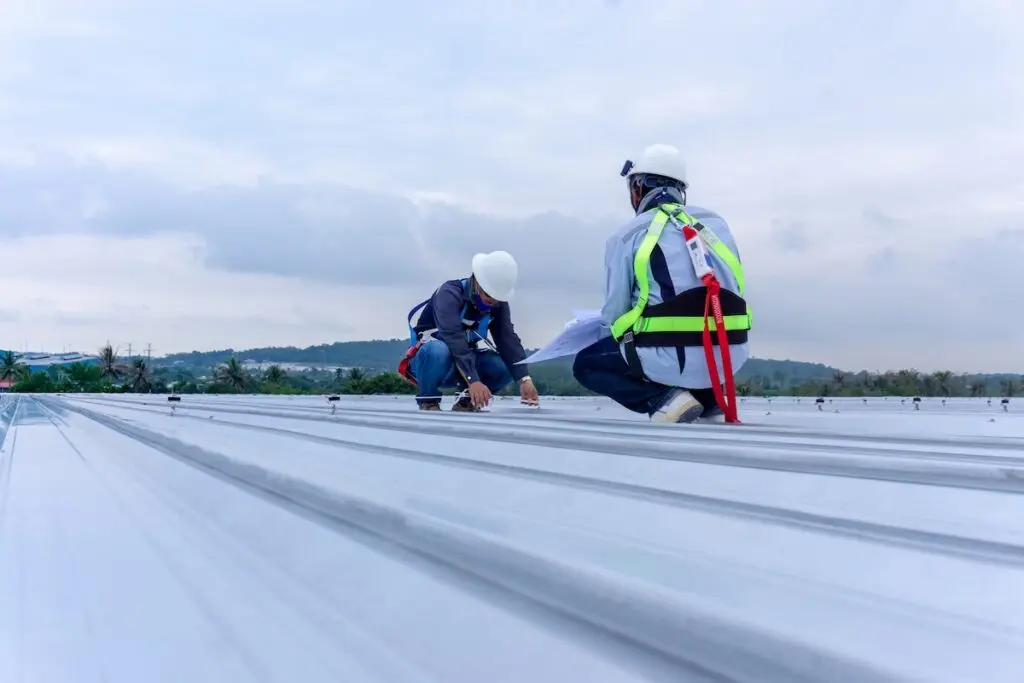 Two construction workers wearing safety harnesses and helmets are kneeling on a metal rooftop, performing a commercial roof inspection together under a cloudy sky with distant hills in the background.