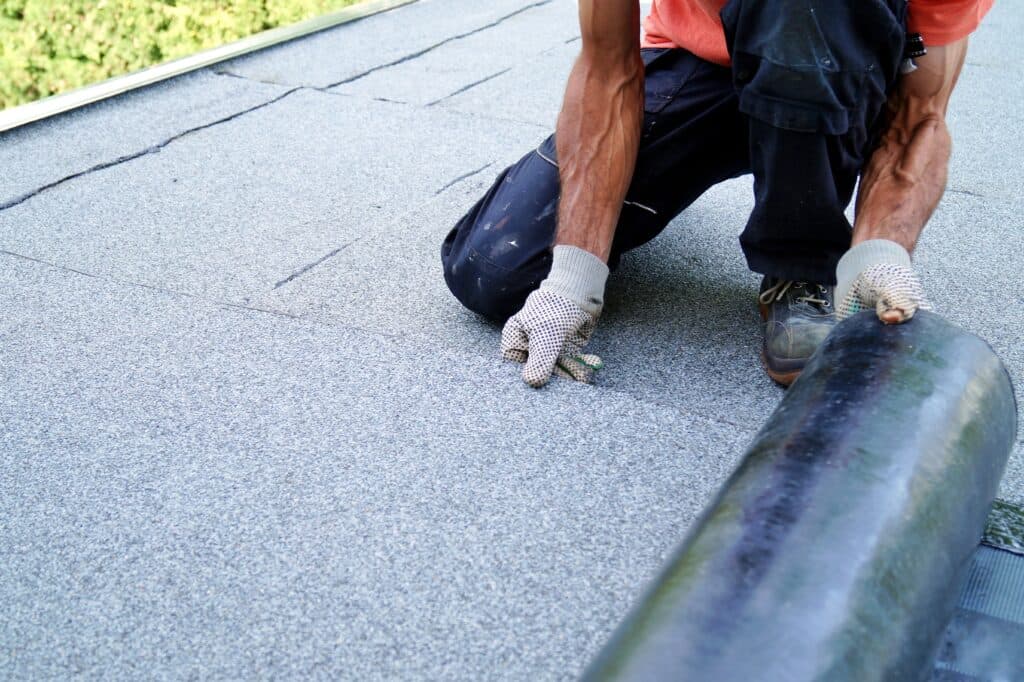 commercial flat roof repair A person wearing gloves kneels on a flat roof, unrolling black roofing material while installing or repairing the surface.