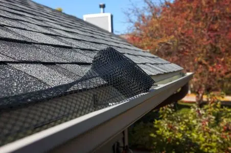 gutter guard installation services A close-up of a house roof shows a black mesh gutter guard being installed, with autumn trees in the background.