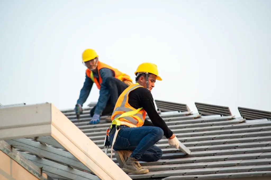 Two construction workers wearing safety vests and yellow helmets are working on a sloped roof, possibly answering how long does it take to replace a roof as they install or inspect metal roofing panels under a clear sky.