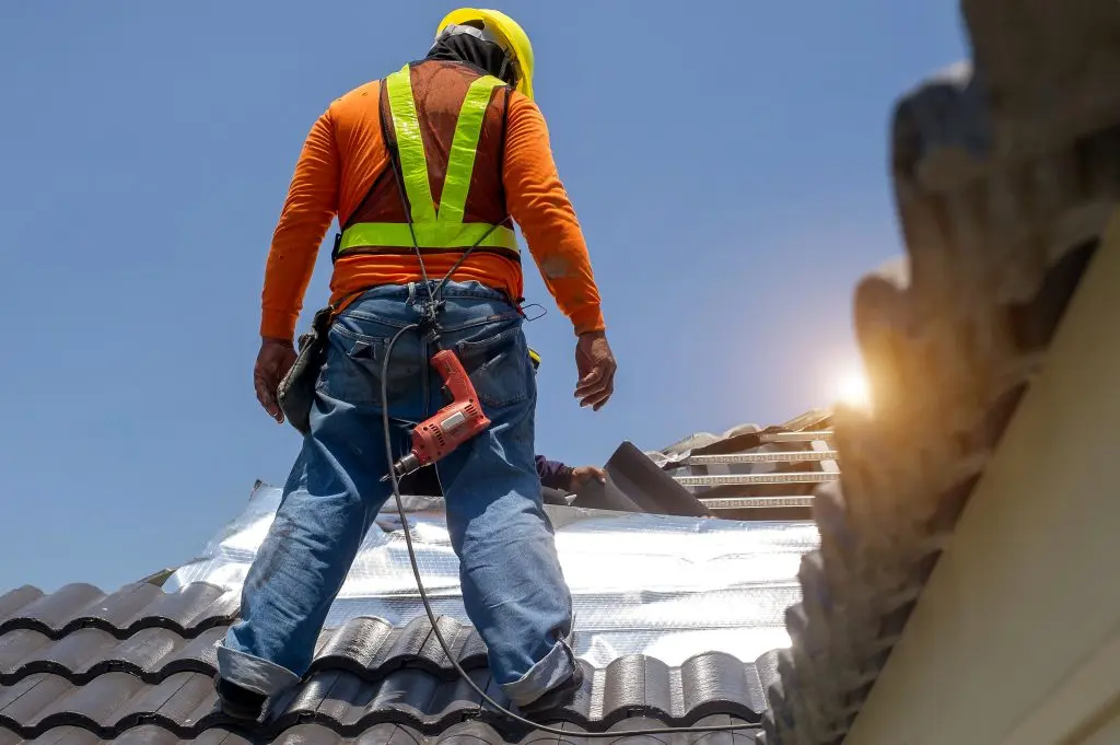 A construction worker in a safety vest and helmet stands on a tiled roof, using a power drill, with sunlight in the background.