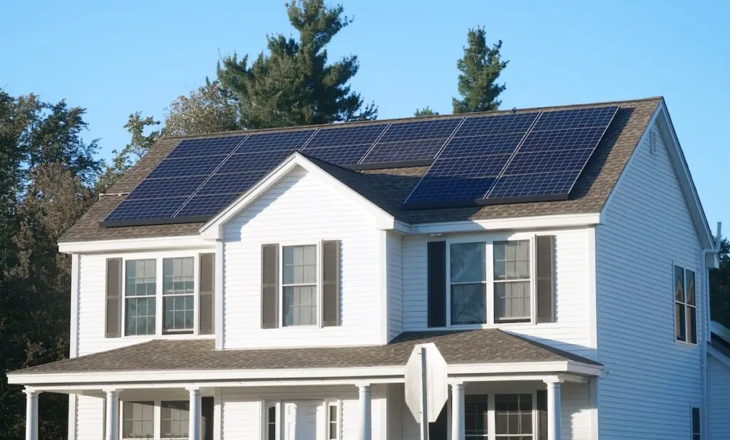 A white two-story house with gray shutters features solar panels on its roof. There are trees in the background under a clear blue sky. A stop sign is partially visible in front of the house.
