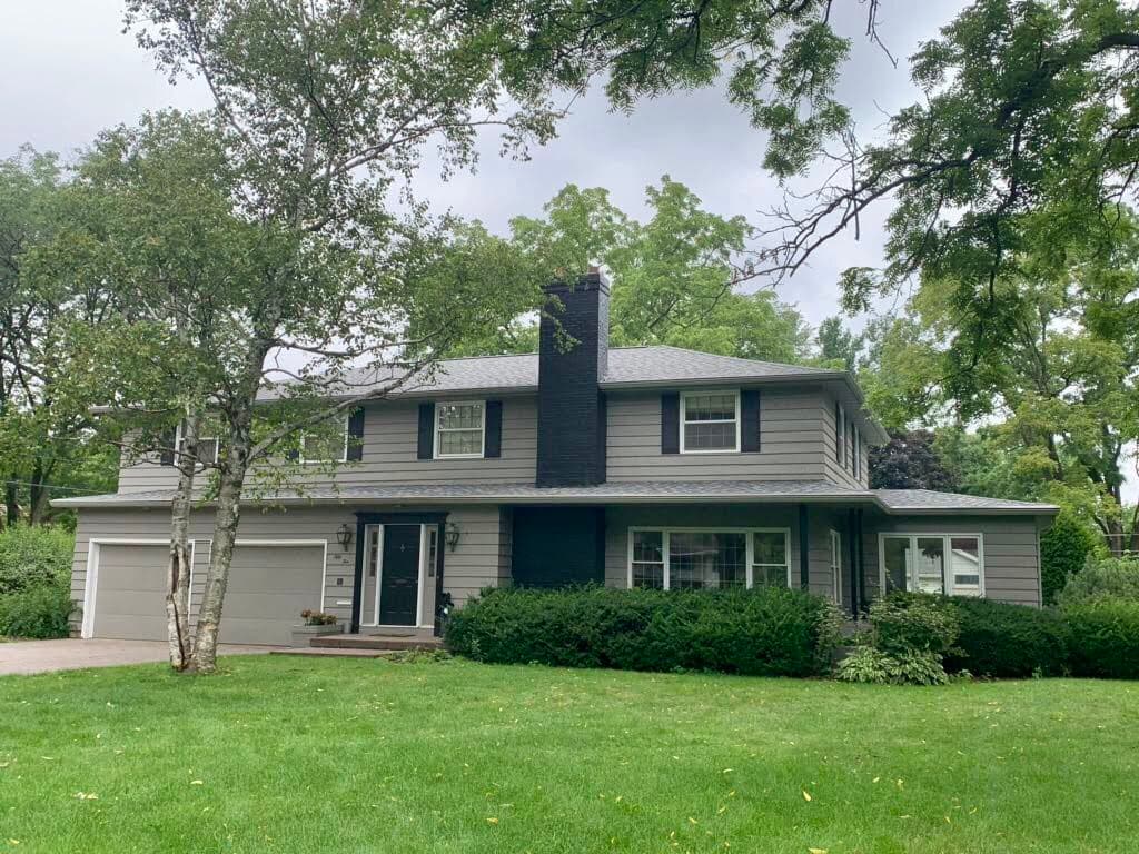 Two-story gray house with white trim, black chimney, and attached garage showcases quality roofing. Located in Pewaukee, it features a large front lawn, surrounded by lush green trees and bushes on a cloudy day.