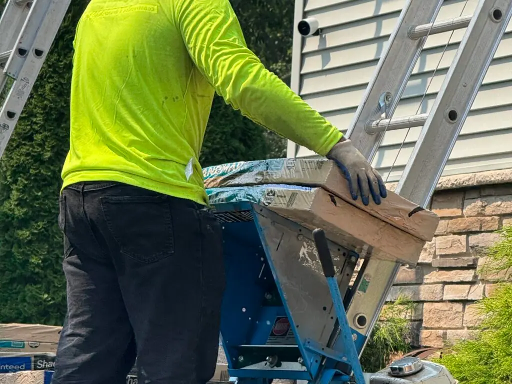A worker in a neon yellow shirt and black pants stands by a blue construction machine near a stack of tile boxes and a stone wall, holding a piece of wood while wearing gloves.