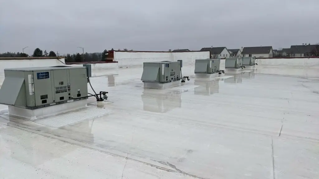 roofing companies verona wi Several large HVAC units are installed on a flat, white commercial rooftop on a cloudy day, with some neighboring buildings visible in the background. The roof surface appears wet and reflective.
