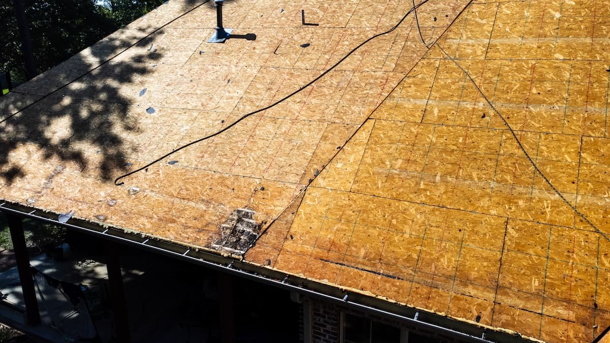 A pitched roof is shown with a wooden sheathing, partially covered by shadows. A chimney pipe and black cable are visible. The roof is bordered by a gutter at the bottom. The surface appears unfinished, awaiting additional layers.