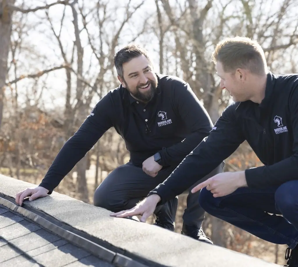 Two men in dark clothing smile and talk while inspecting the edge of a roof on a sunny day, with leafless trees visible in the background.