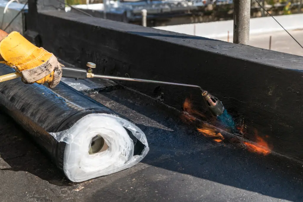 types of commercial roofing systems A worker wearing a yellow glove uses a blowtorch to heat and apply a roll of waterproofing membrane onto a flat rooftop during construction.