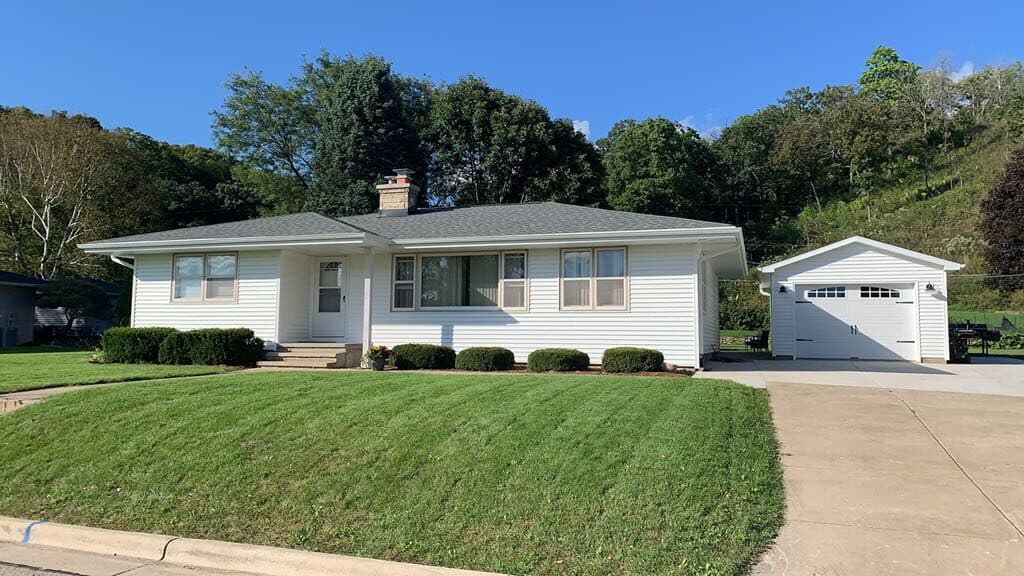 A single-story white house with gray roof, neatly trimmed bushes, and green lawn sits next to a detached white garage with a driveway on a sunny day—showcasing top-quality roofing Milwaukee homeowners appreciate. Trees and a small hill are in the background.