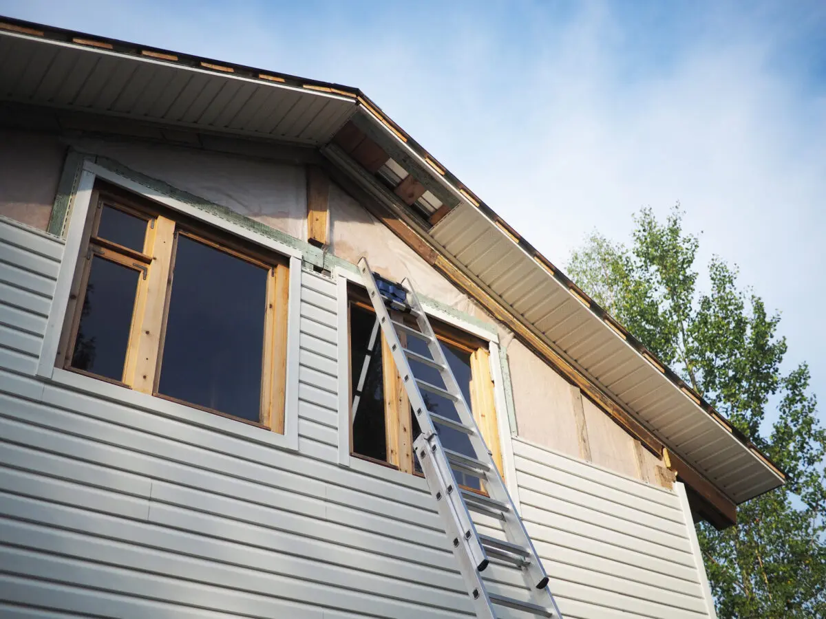fiber cement siding installation A metal ladder is propped up against the side of a house with partially finished siding and exposed wooden framing around the upper windows, under a blue sky with some trees visible nearby.