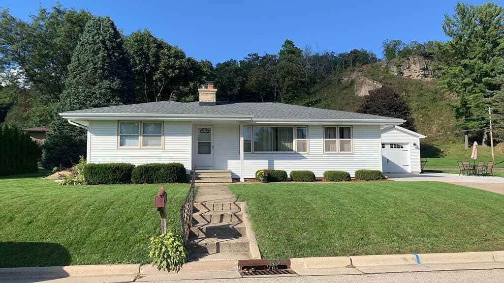 A single-story white house with a gray roof, front porch, and attached garage sits on a well-manicured lawn. This charming home showcases quality roofing Brookfield residents can trust, nestled among trees and rocky hills under a clear blue sky.