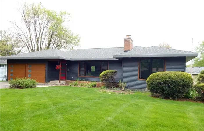 A modern single-story house with dark blue siding and a contrasting wooden garage door. The front yard features a neatly maintained lawn, landscaped shrubs, and a few trees in the background. Large windows and a red door add distinctive accents.