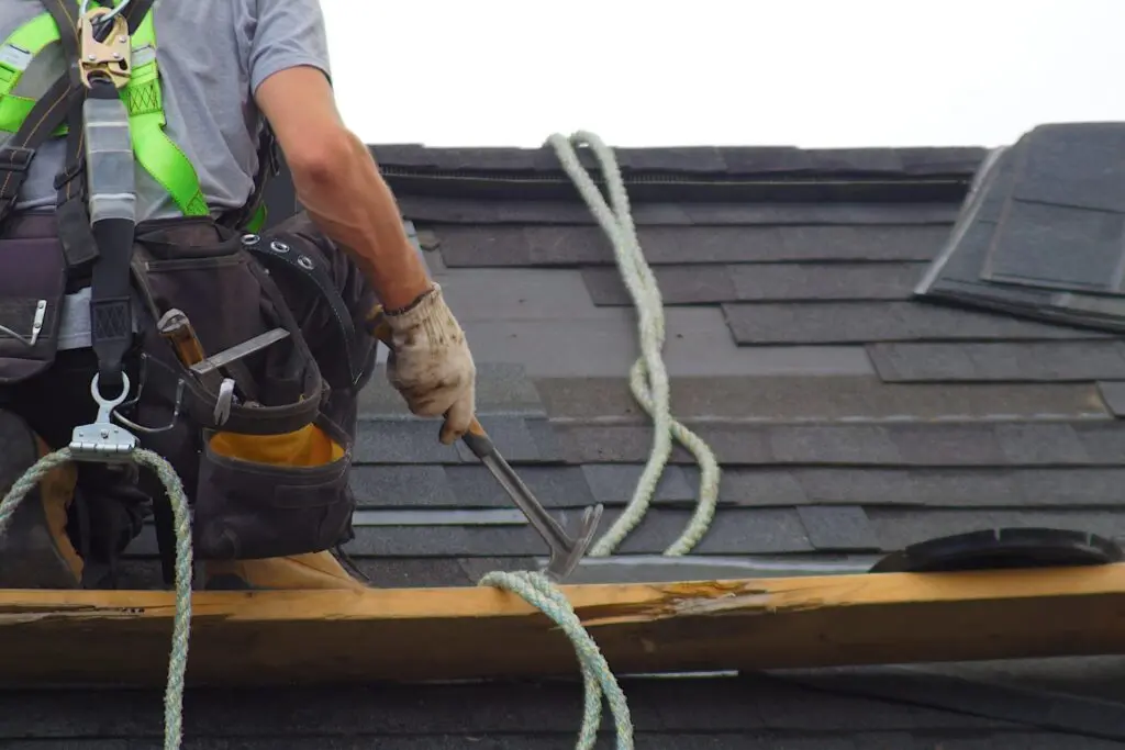 A construction worker wearing a safety harness and gloves uses a hammer while working on a shingled roof. Roofing materials and ropes are visible on the roof surface.