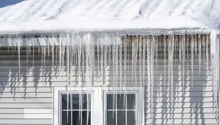 ice and water shield roof Long icicles hang from the snowy roof edge above a white house with vertical siding and glass doors, casting striped shadows on the wall in bright sunlight.