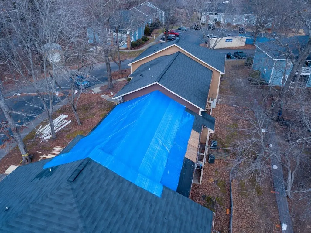 Aerial view of a house with a large blue tarp covering part of its roof. The surrounding area is filled with trees without leaves, suggesting a winter setting. Nearby, other houses are visible, and scattered wooden planks lie on the ground.